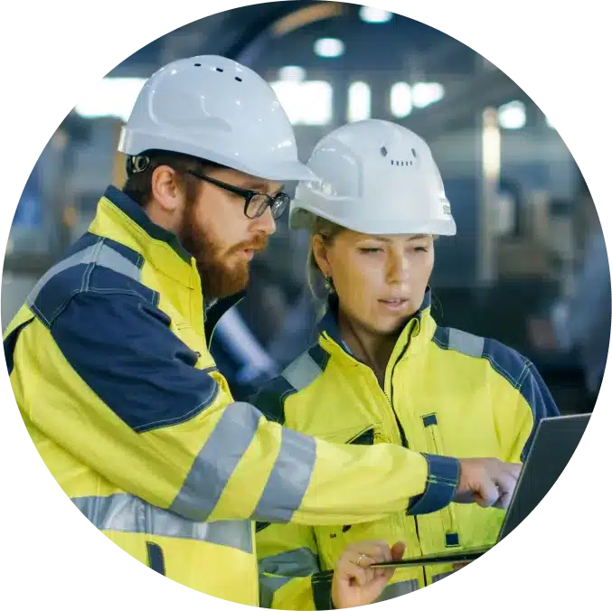 Two engineers in safety gear discuss over a laptop in an industrial setting.