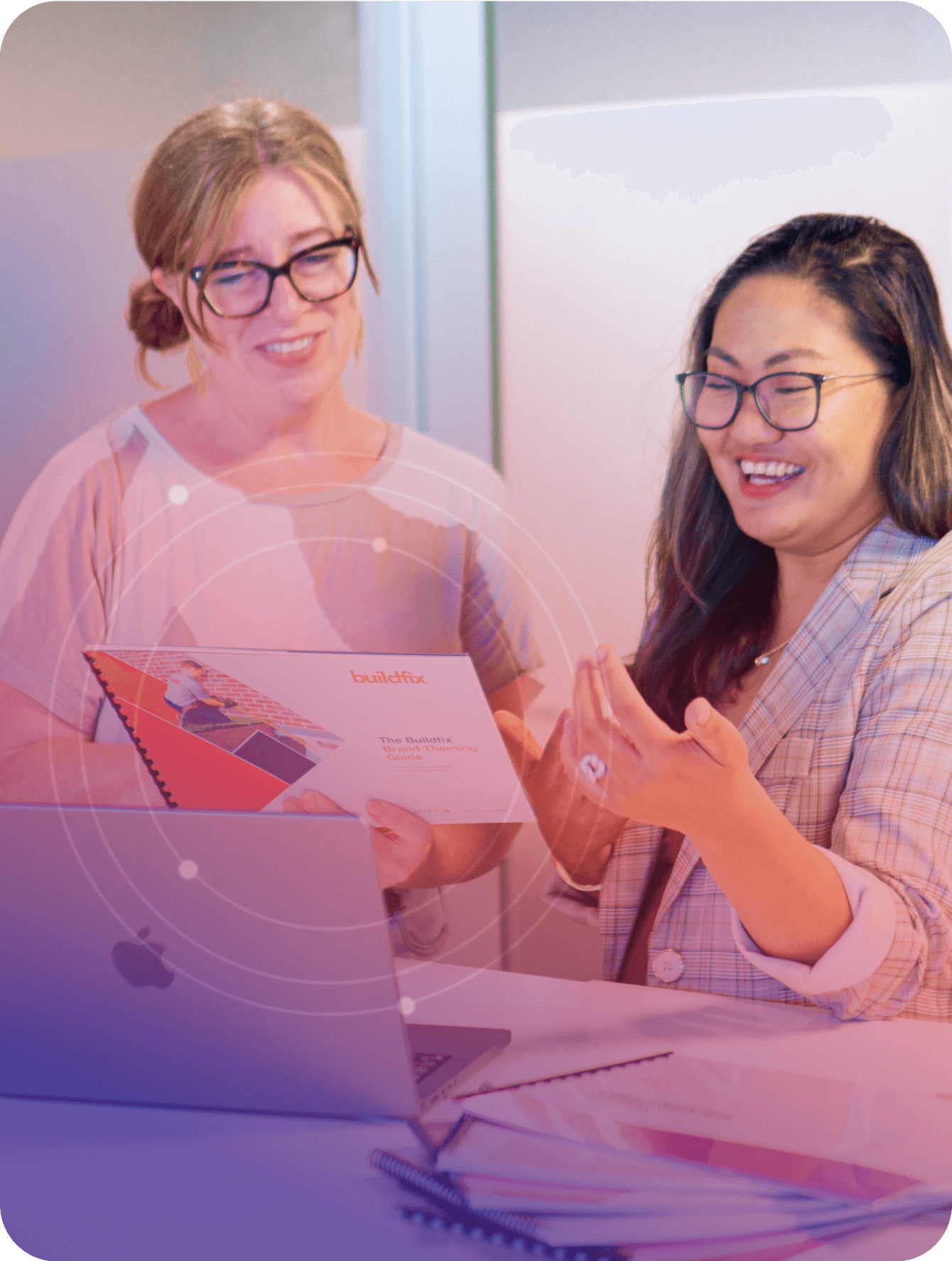 Two women in a casual office setting are smiling and discussing a booklet labeled "buildfix," which includes their capability statement, with a laptop open on the desk in front of them.