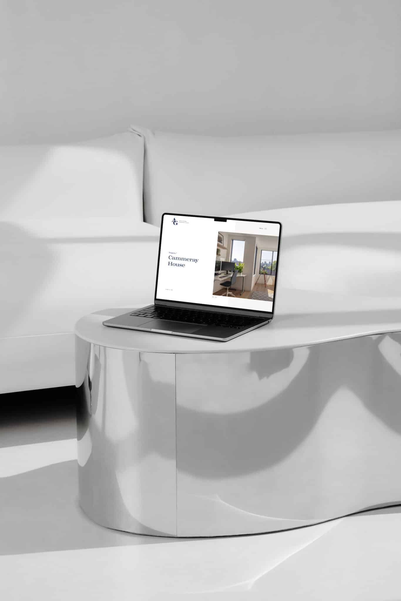 A sleek A&G laptop sits on a chrome coffee table in a minimalist room with a white couch. Its screen displays the "Commencing Future" website, showcasing an image of an elegant interior space.