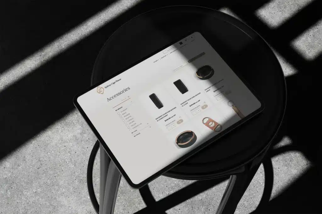 A tablet showcasing an online store page for accessories, testament to expert eCom Web Development, rests on a black stool, bathed in shadows and sunlight on the concrete floor.
