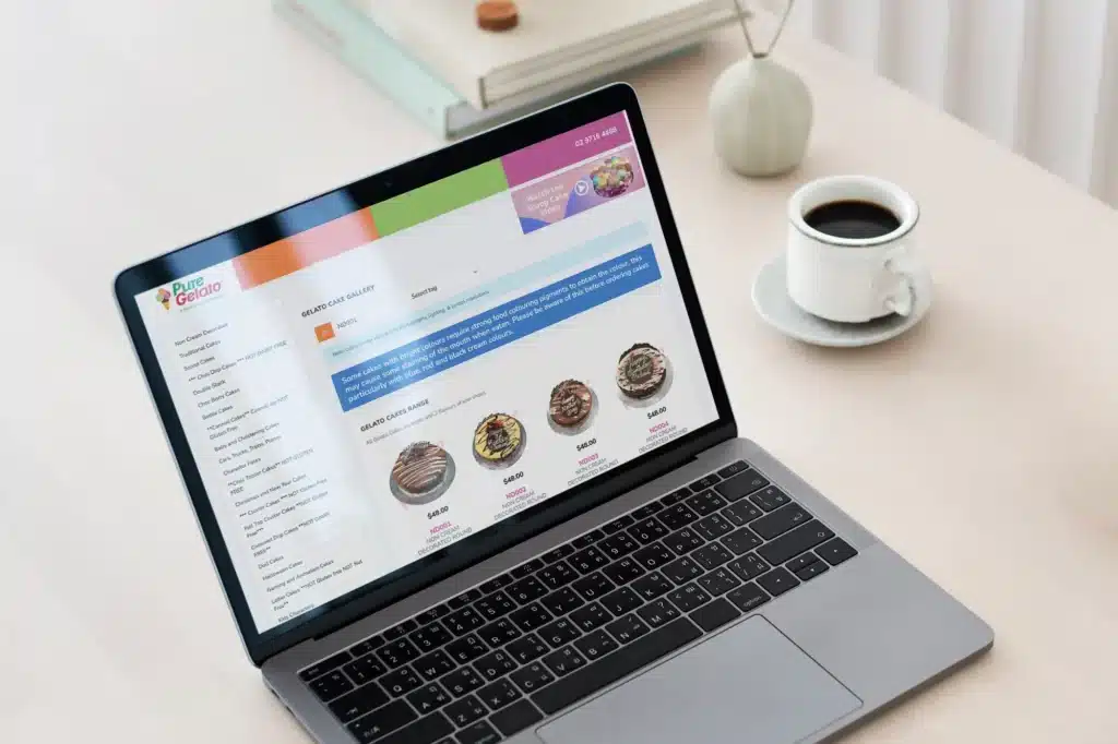 A laptop on a table displays a webpage with an online grocery store interface, looking sleek and organized like a digital portfolio. A cup of coffee and a notebook are nearby, setting the scene for productivity.