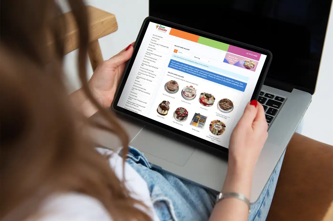 A person is seated at a table with a laptop, holding a tablet that showcases cake images and descriptions on an eCommerce webpage, highlighting the intersection of digital design and web development.