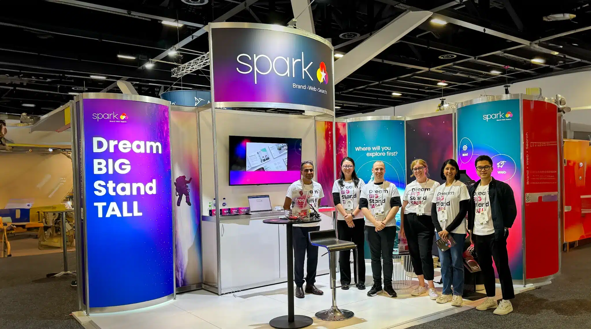 A group of six people stands proudly in front of a colorful exhibition booth featuring "Dream Big Stand Tall" and "spark" branding. The booth, showcasing their main services, includes a screen, posters, and a small table with stools—creating an inviting page in any SEO strategy.