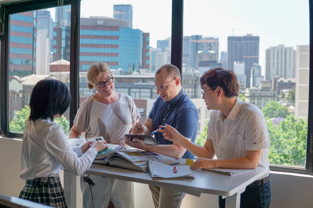 Four people stand around a table with books and papers, deep in discussion, in a brightly lit office with city buildings visible through large windows—a scene reminiscent of a Spark Interact team preparing for the 2025 Australian Web Award.