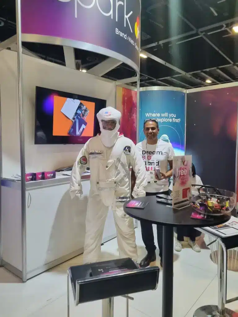 A person in a white astronaut suit stands at a tech exhibition booth, engaging in business networking, while another casually dressed individual browses promotional materials on display.