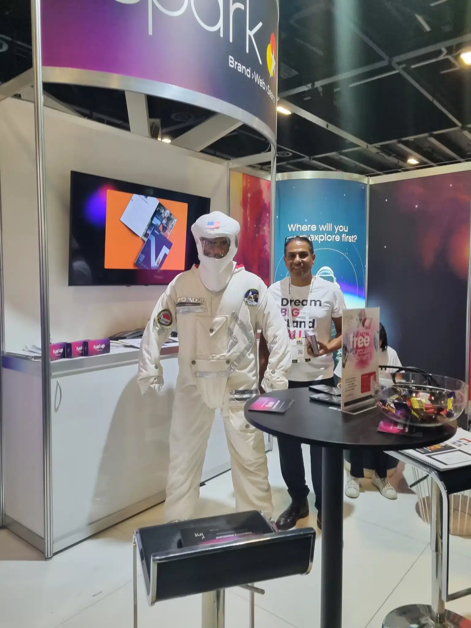 A person in a white astronaut suit stands at a tech exhibition booth, engaging in business networking, while another casually dressed individual browses promotional materials on display.