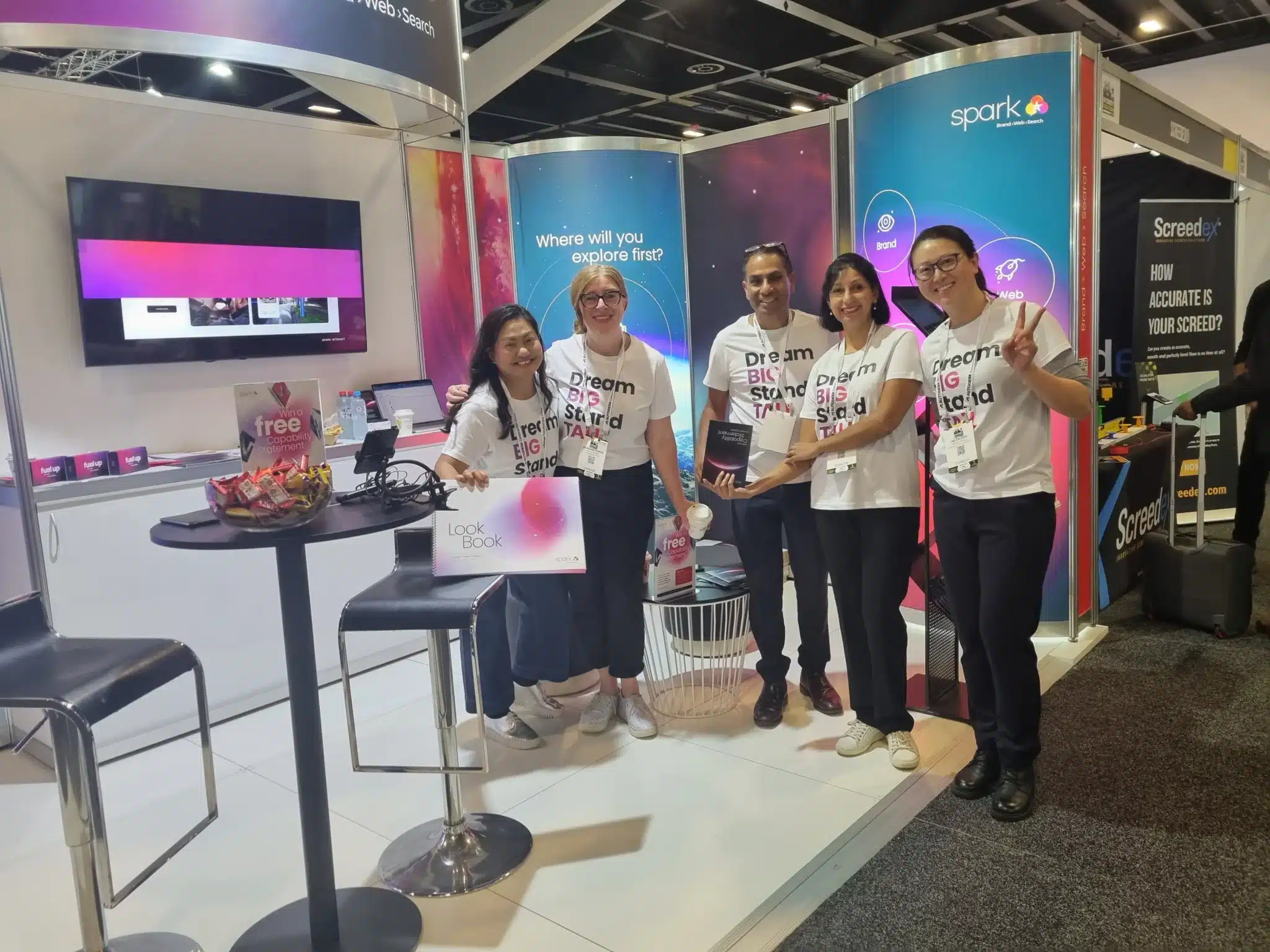 Spark team wearing matching t-shirts stand and smile at a technology expo booth, showcasing promo materials and display screens that highlight their successful expo marketing and booth to business strategies.