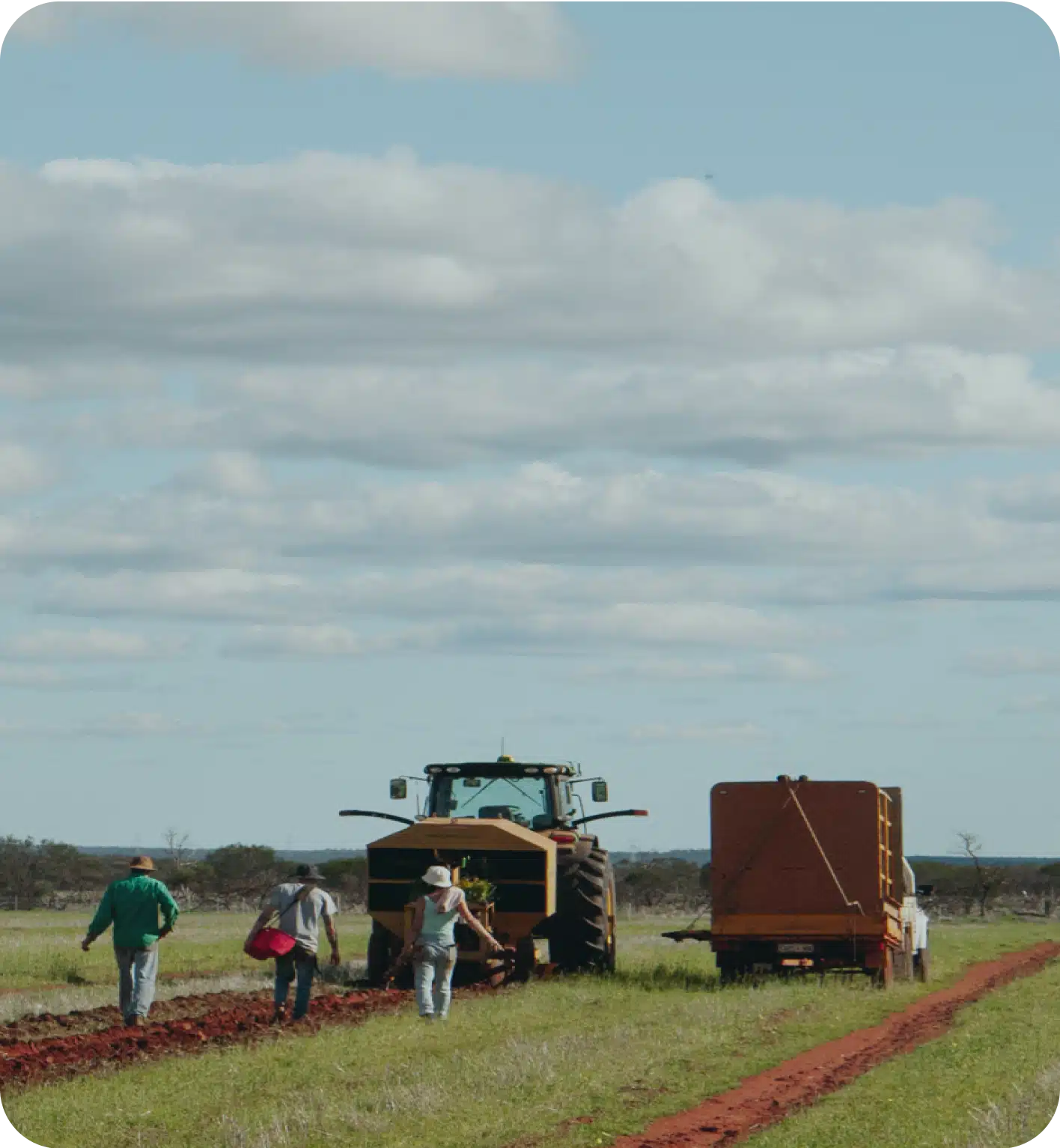 Four people walk on a dirt path beside a tractor and trailer in a grassy field under a partly cloudy sky.