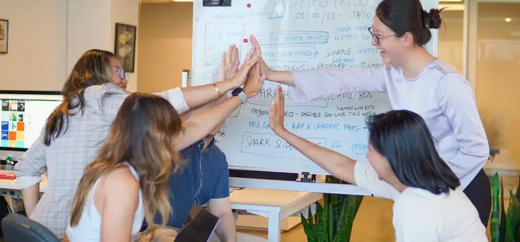 Five people sitting and standing around a whiteboard in an office give each other a group high-five, appearing to celebrate or collaborate on a project.