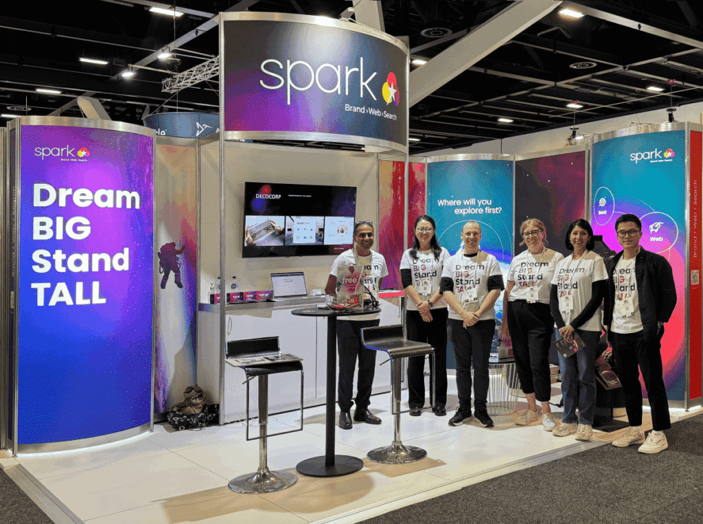Six people stand in front of a trade show booth for "spark," featuring bright signs, a table with chairs, and digital displays that showcase integrated marketing communication and innovative marketing tools.