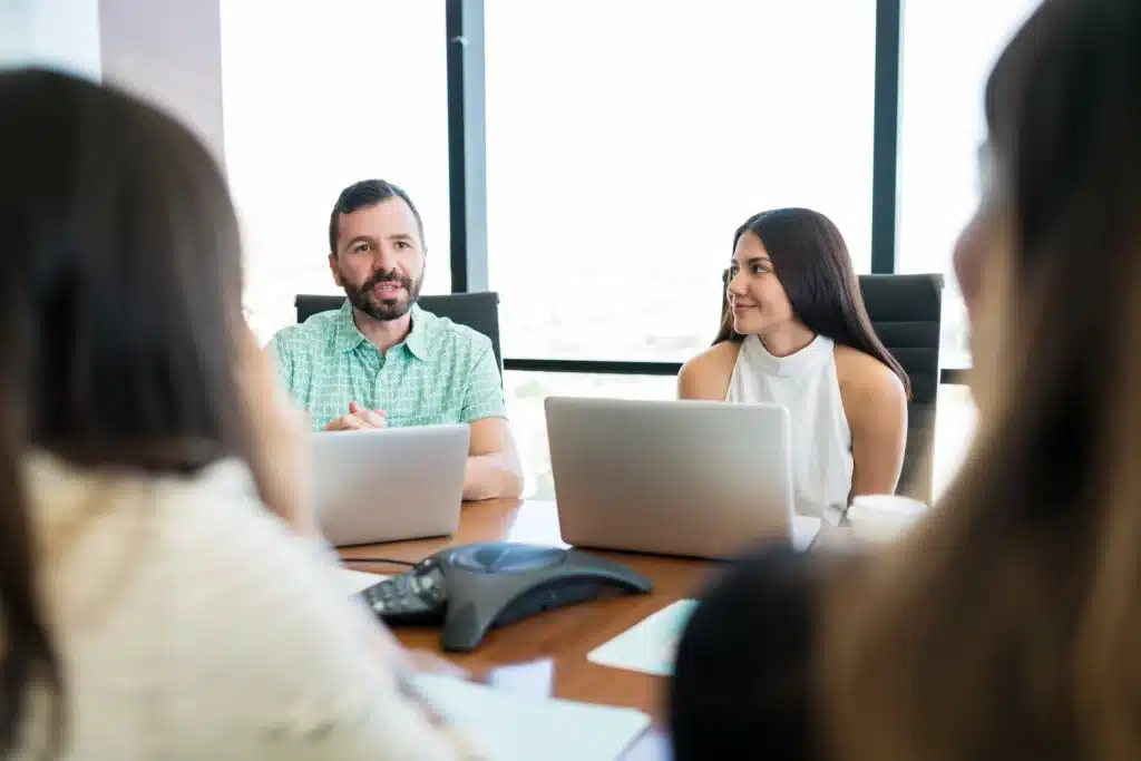 Four people sit around a conference table with laptops, engaged in a meeting focused on AI Optimisation. The man and woman in focus appear to be speaking and listening, while a speakerphone rests on the table.