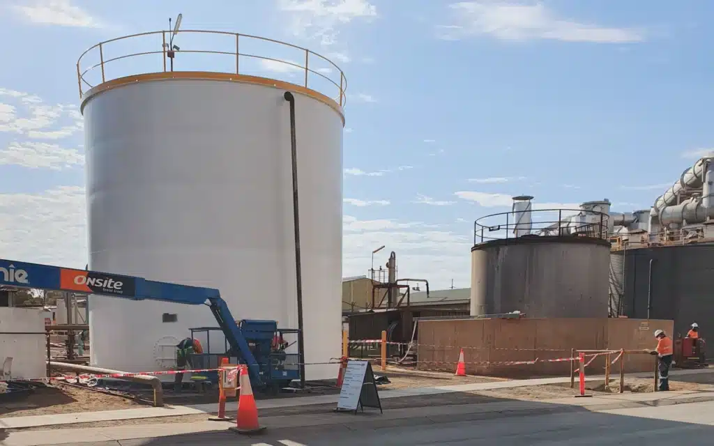 A large white industrial storage tank is being serviced by workers, including a BHP Veteran, using a blue lift and surrounded by safety cones and barriers at an industrial site in the Goldfields under a partly cloudy sky.