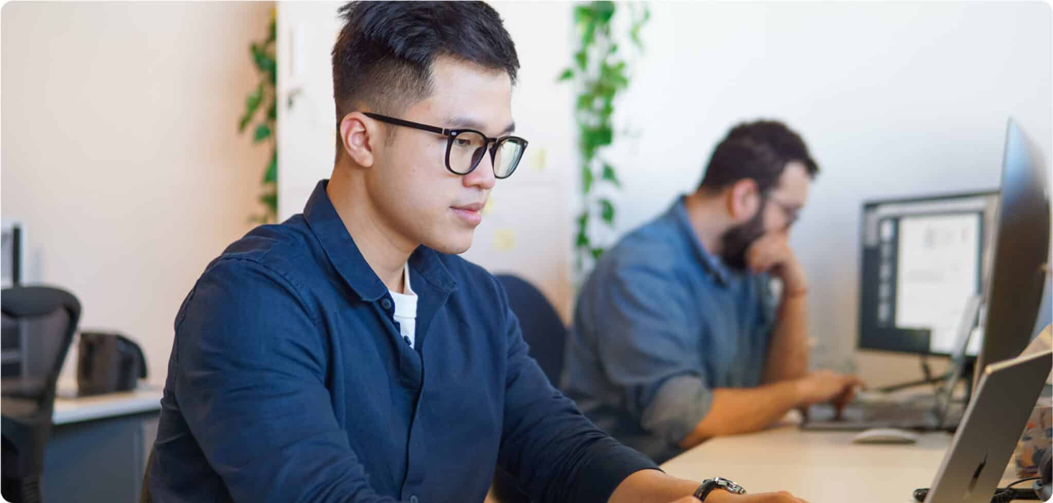 Two men work at desks with computers in a modern office space, reflecting a sleek 2025 design. The man in the foreground is typing on a laptop, while the man in the background is focused on his desktop monitor, updating the Home Page.