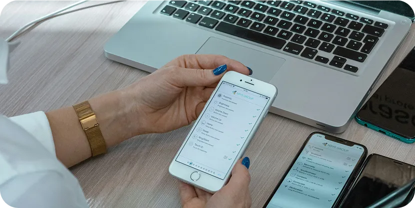 A person holding a smartphone with a Website Maintenance 2025 checklist app open, next to an open laptop and another phone on a wooden desk.
