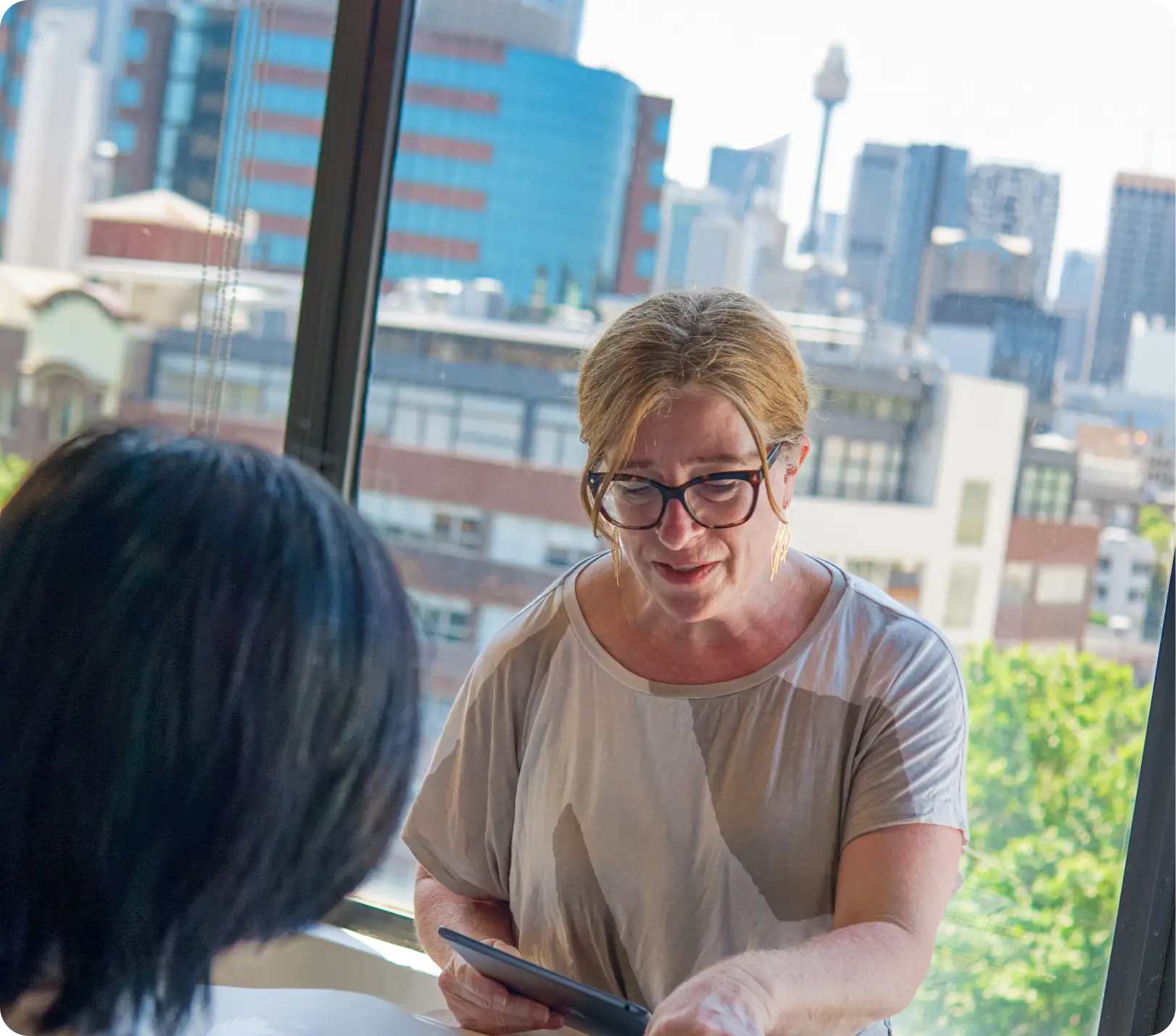 Two people sit by a window in an office with a cityscape in the background as one holds a tablet and discusses updates for the 2025 Home Page.