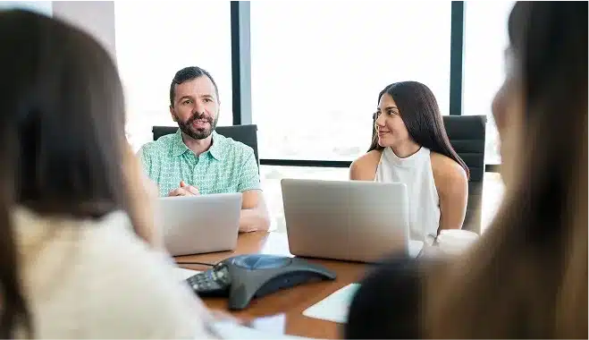 A man and a woman sit at a conference table with laptops, discussing Web Services in a 2025 strategy meeting with two other people in a modern office.