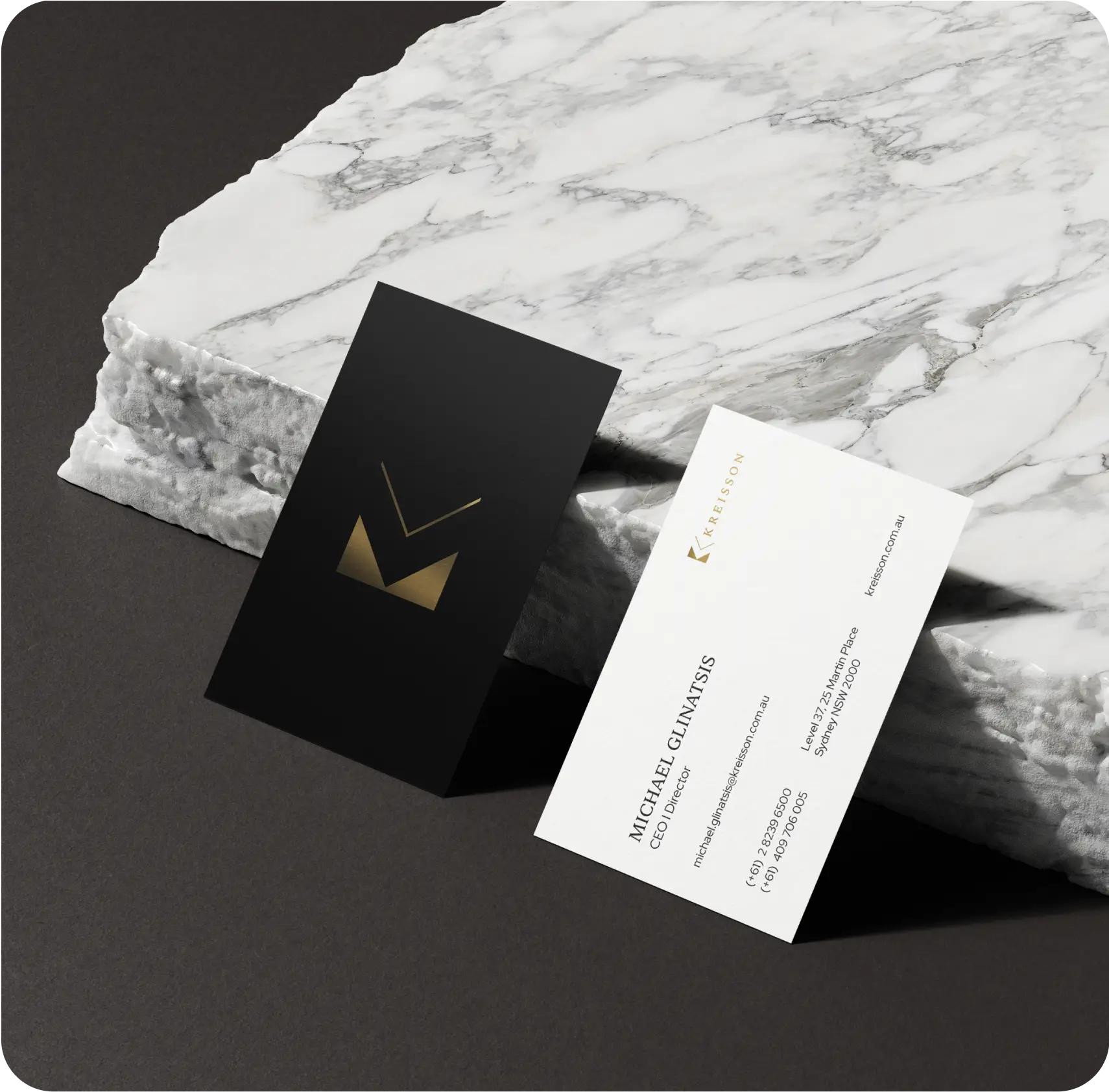 Two business cards, one black with a gold geometric logo and one white with company and contact details, displayed on a white marble slab.