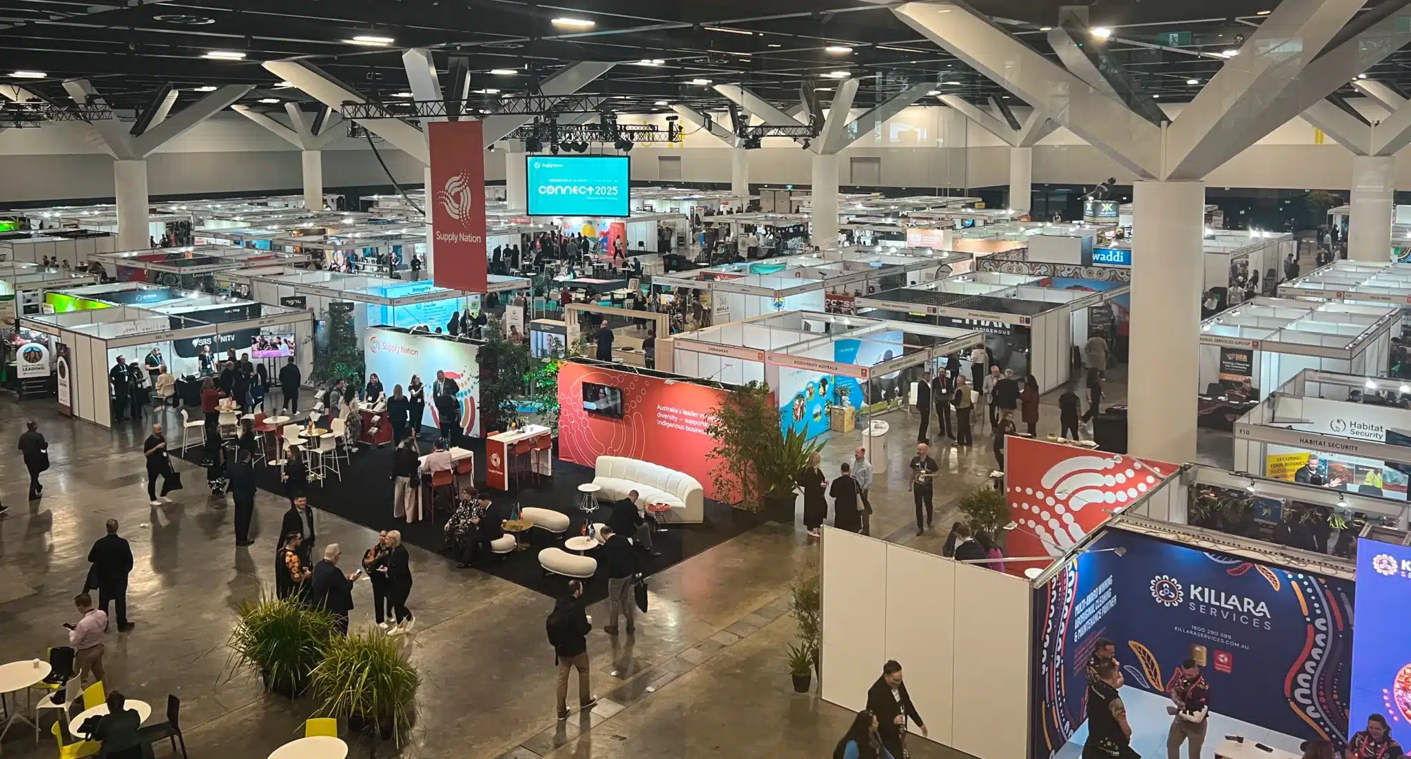 Wide view of a busy indoor convention center at Supply Nation Connect 2025, featuring numerous exhibition booths, information displays, banners, and people walking or interacting.