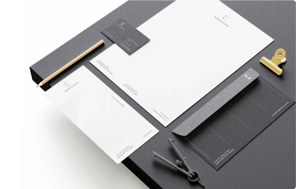 A set of branded stationery items including letterhead, envelope, business card, and notepad, arranged neatly on a dark surface with a gold pen, binder clip, and drawing compass.