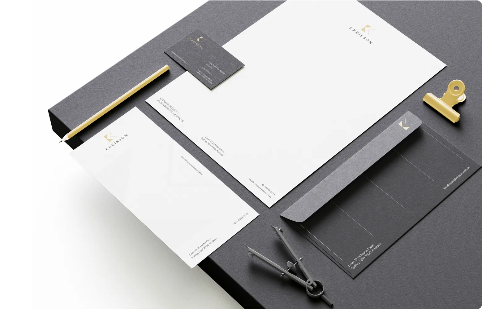 A set of branded stationery items including letterhead, envelope, business card, and notepad, arranged neatly on a dark surface with a gold pen, binder clip, and drawing compass.