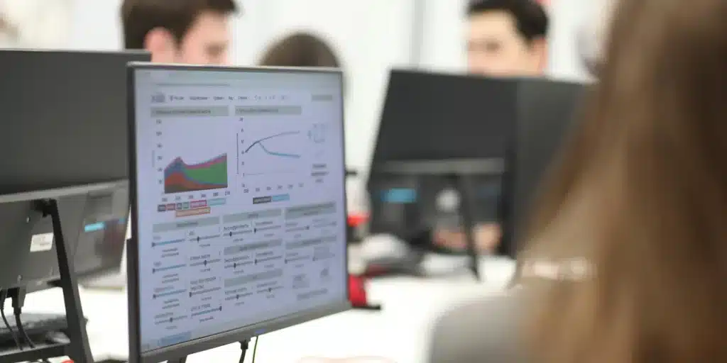 A computer monitor displays data charts and graphs in an office setting, highlighting analytics for a Search Service in 2025, with people working at desks in the background.