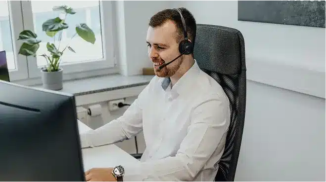 A man wearing a headset and white shirt sits at a desk in front of a computer monitor, working with Web Services in an office environment, preparing for the demands of 2025.