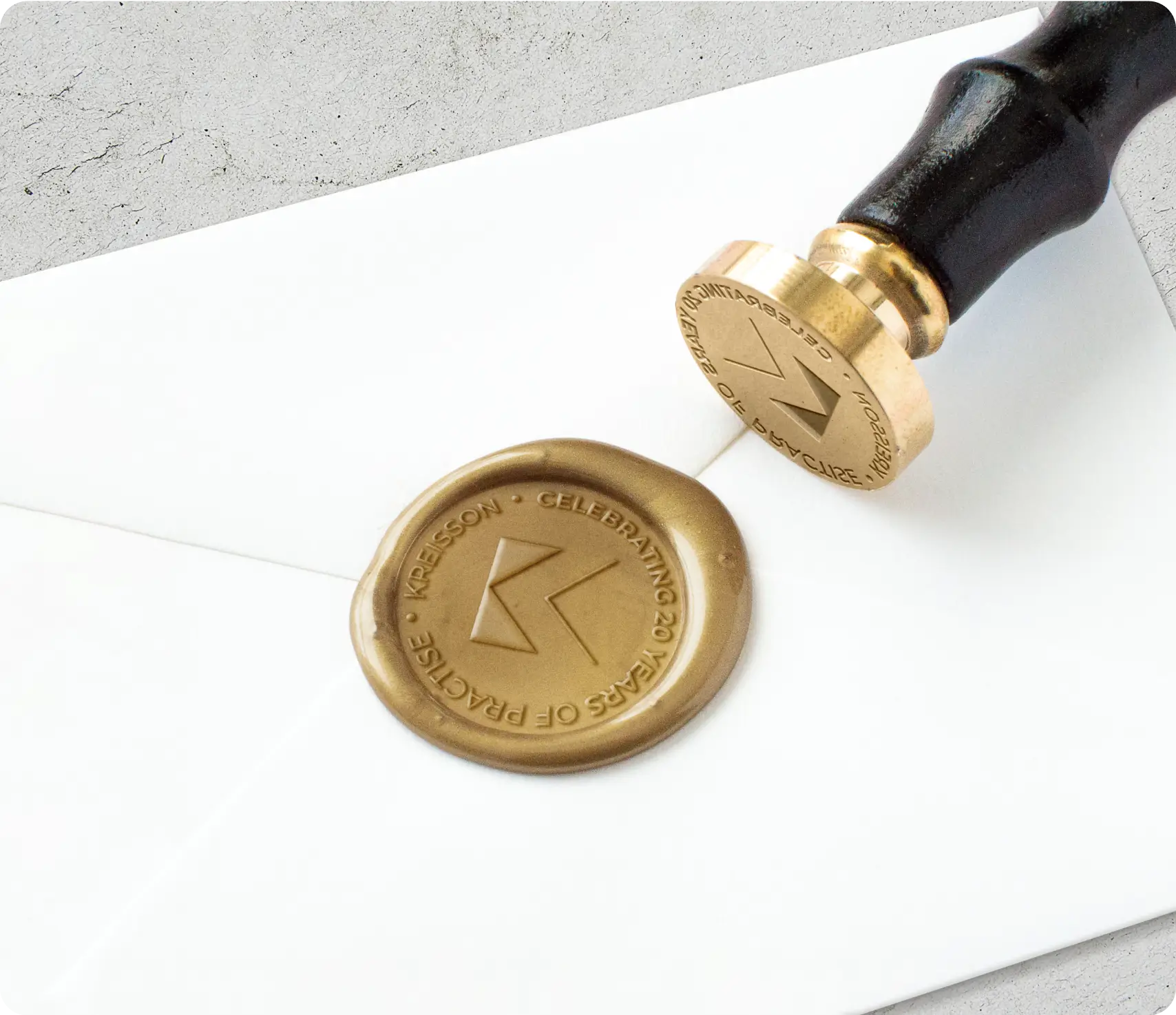 A golden wax seal stamped on a white envelope, with a metal stamp resting nearby; the seal features a "K" and circular text.