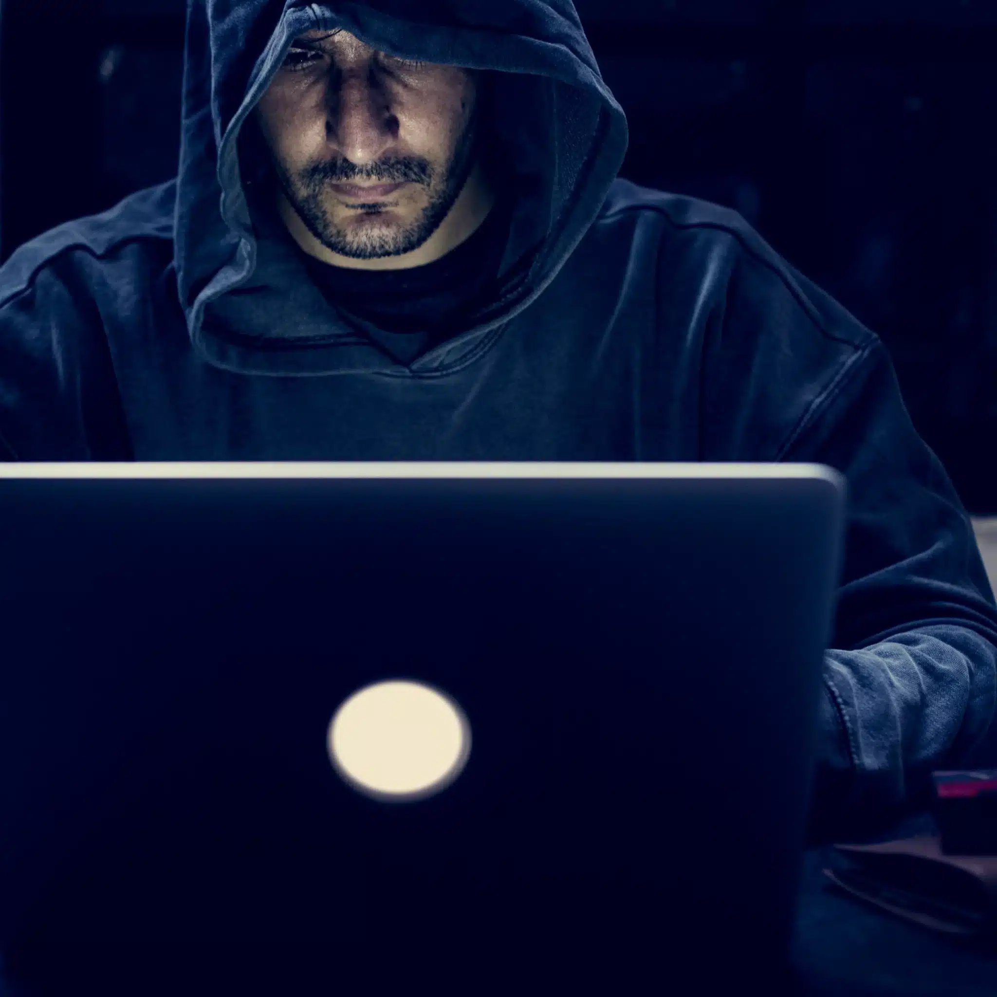 A person wearing a dark hoodie sits in front of a laptop, the screen lighting up their face as they work on a website redesign in a dimly lit environment.