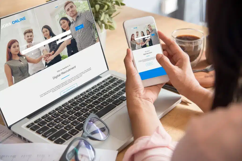 A person in Australia holds a smartphone displaying a jobs website that matches the one on their laptop, alongside glasses and coffee on the desk—perfect for following a website checklist or planning to redesign your website.