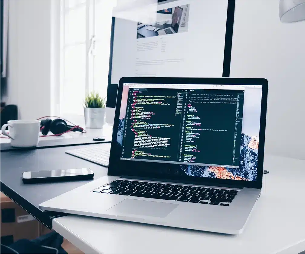 A laptop displaying code sits on a white desk next to a smartphone, headphones, a coffee cup, and an external monitor showing Web Services for 2025.