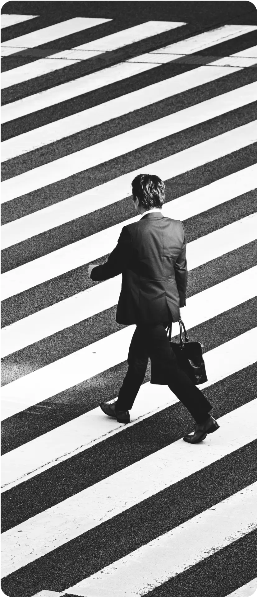 A person in a suit carrying a bag walks across a crosswalk with bold, diagonal white stripes.