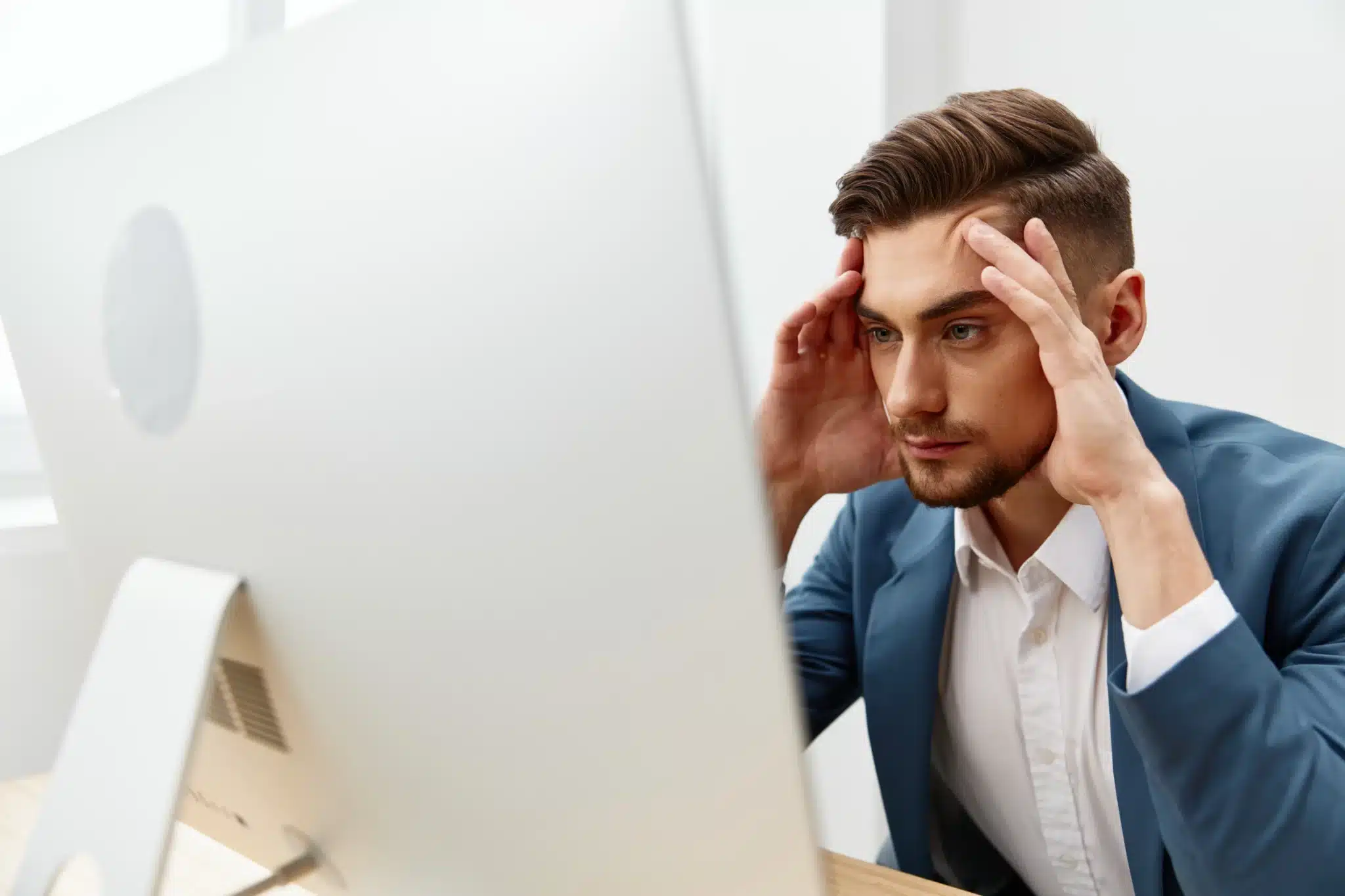 A man in a suit sits at a desk, looking at a computer screen with his hands on his temples, appearing focused or stressed—perhaps weighing options like Basic Web Hosting or Managed Hosting, as many smart business owners do.