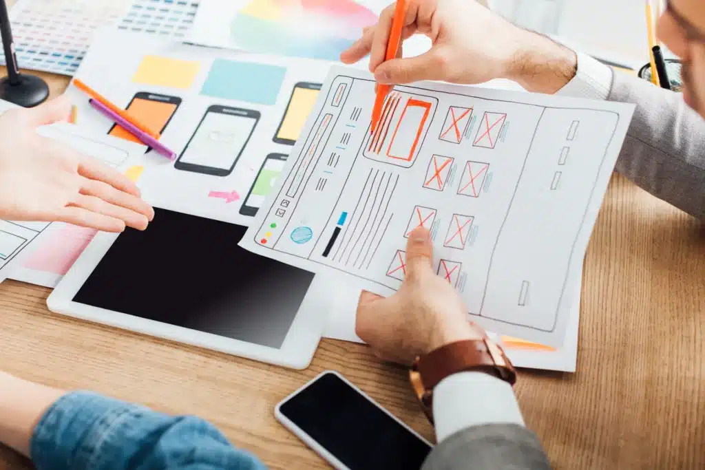 Two people in Australia discuss and review website wireframes and designs on printed paper, with a tablet, smartphone, and more design materials on the desk—perfect inspiration to redesign your website using a free checklist.