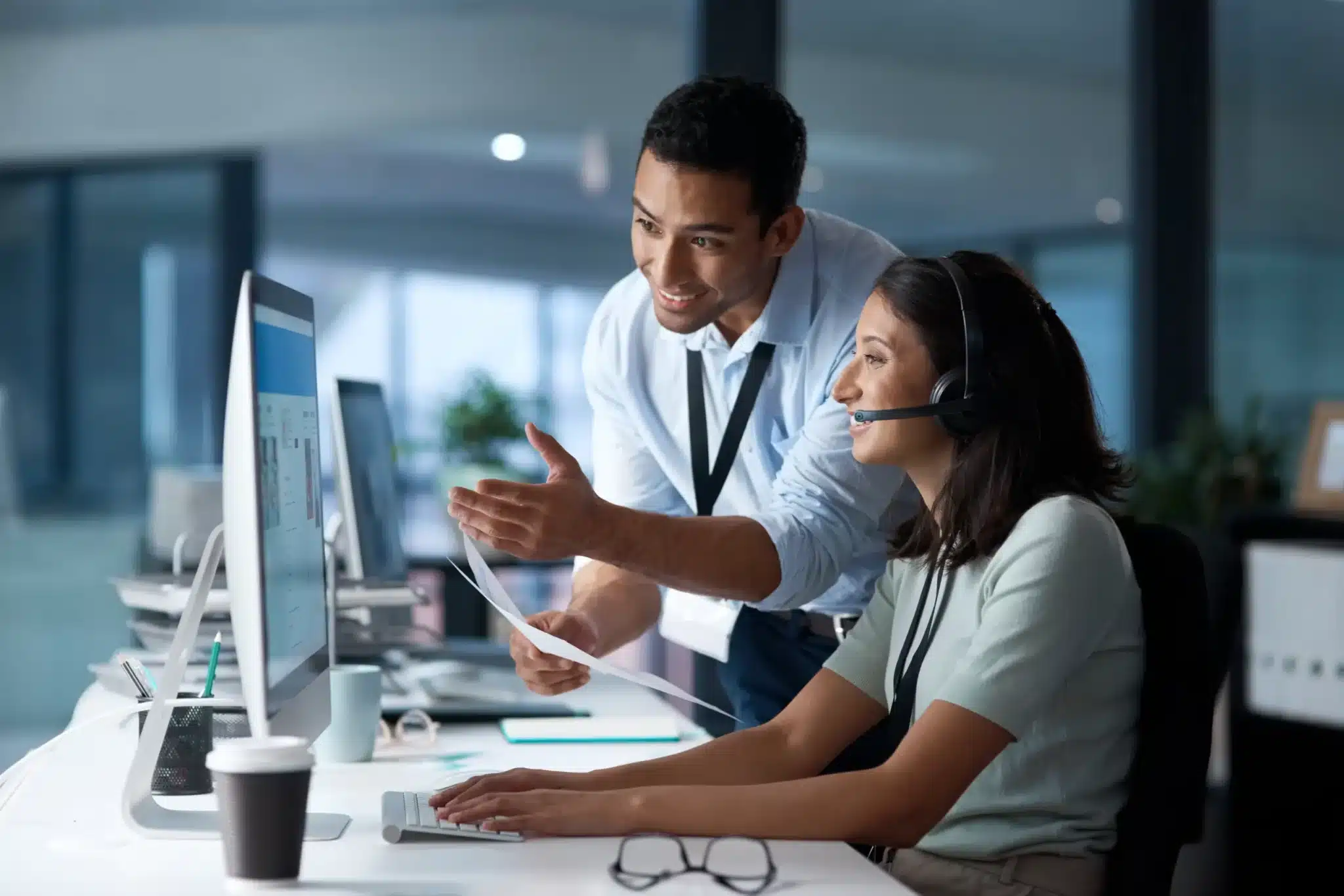 Two office workers at a desk, one seated wearing a headset looking at a computer, the other standing and pointing at the screen, both appearing engaged in discussion about managed hosting for business owners.