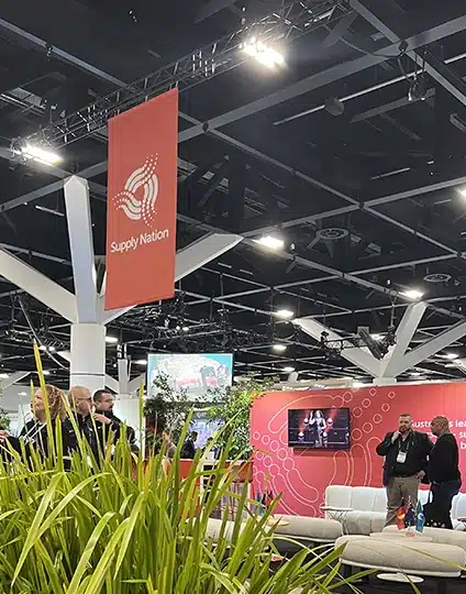 Indoor event space with people gathered near a red "Supply Nation" banner, surrounded by plants and seating, with a TV screen and red backdrop—an inspiring setting for supplier diversity at Supply Nation Connect 2025.