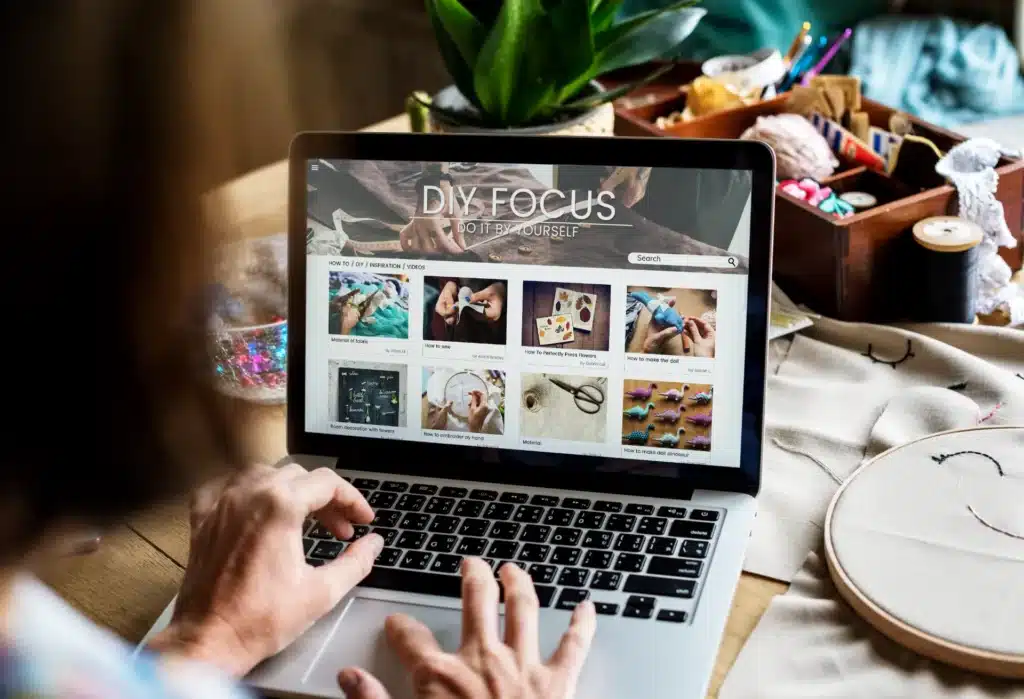Person using a laptop displaying a DIY website with various craft project thumbnails; sewing supplies and embroidery hoop are visible on the table—perfect inspiration if you want to redesign your website in Australia.