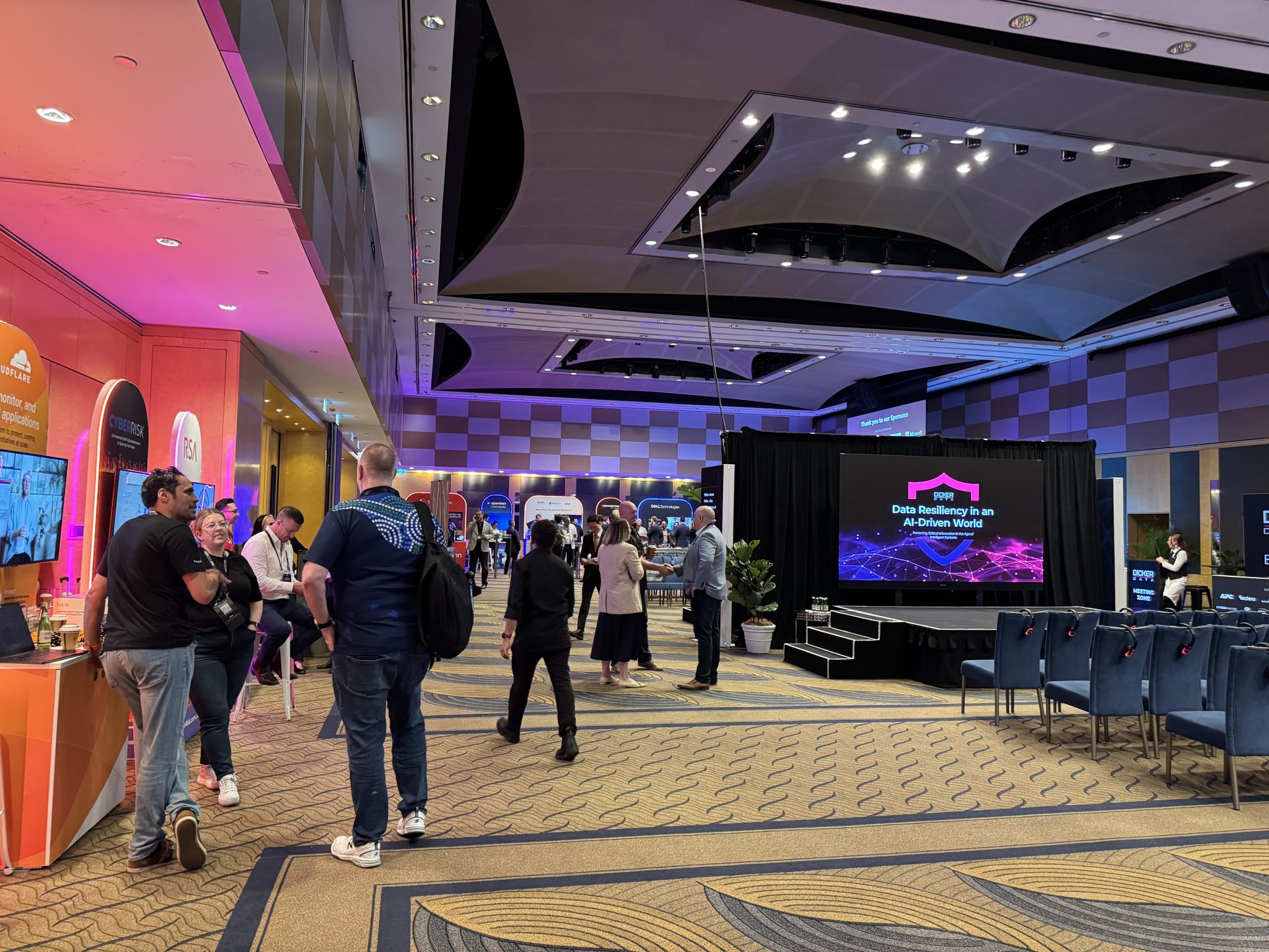 People attend a tech conference in a large indoor venue with booths, a presentation stage, and a display reading "Data Driven Ways to Achieve Value," exploring how cyber resilience offers a competitive advantage.