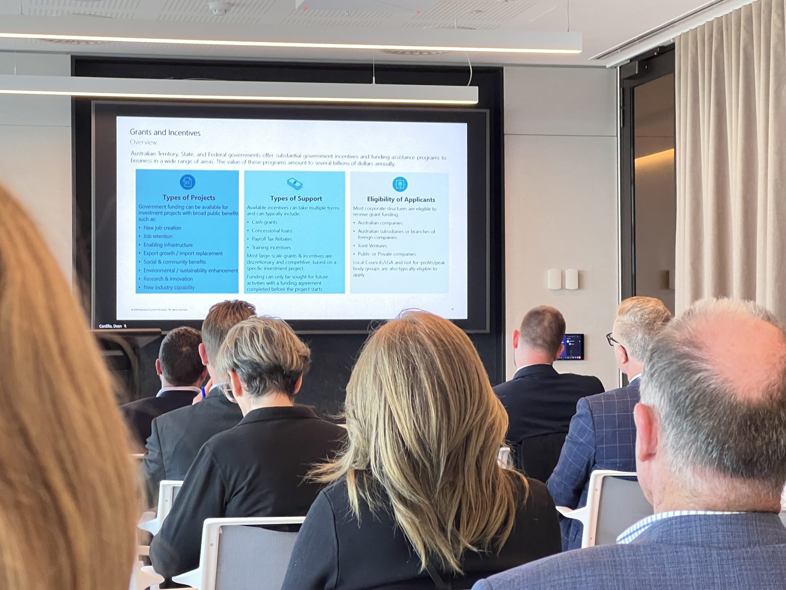 A group of people in business attire attend an AIDN NSW Member Meeting in a conference room; a slide about grants and incentives, presented by Deloitte, is displayed on a large screen.