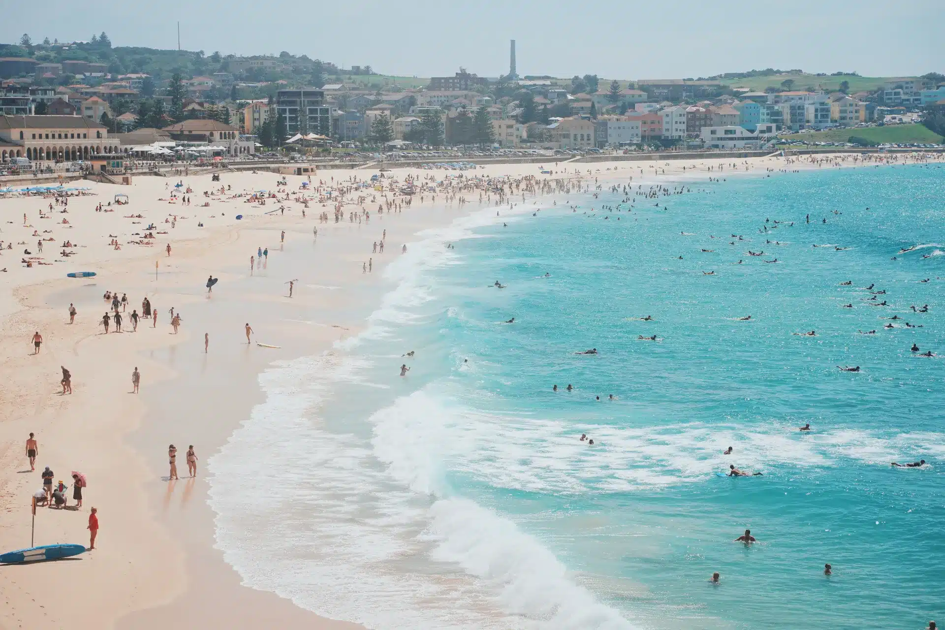 A wide sandy beach in Bondi with people sunbathing on the shore and surfing in the blue ocean; buildings, greenery, and a hint of Web Design Bondi creativity visible in the background.