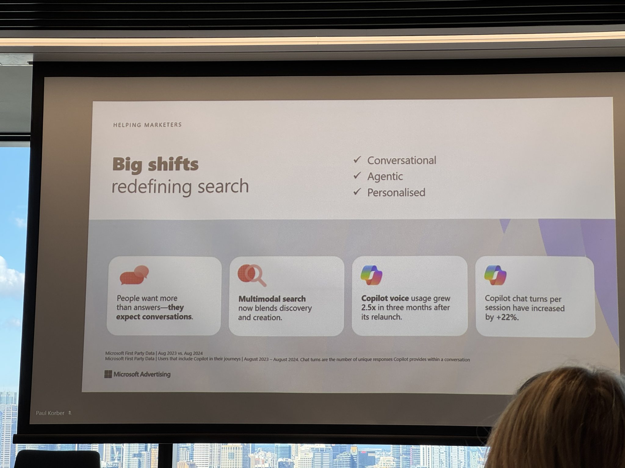 A presentation slide titled "Big shifts redefining search" lists trends in search behavior, with four icons and statistics, highlighting AI-driven changes, and a cityscape visible through the window behind it.