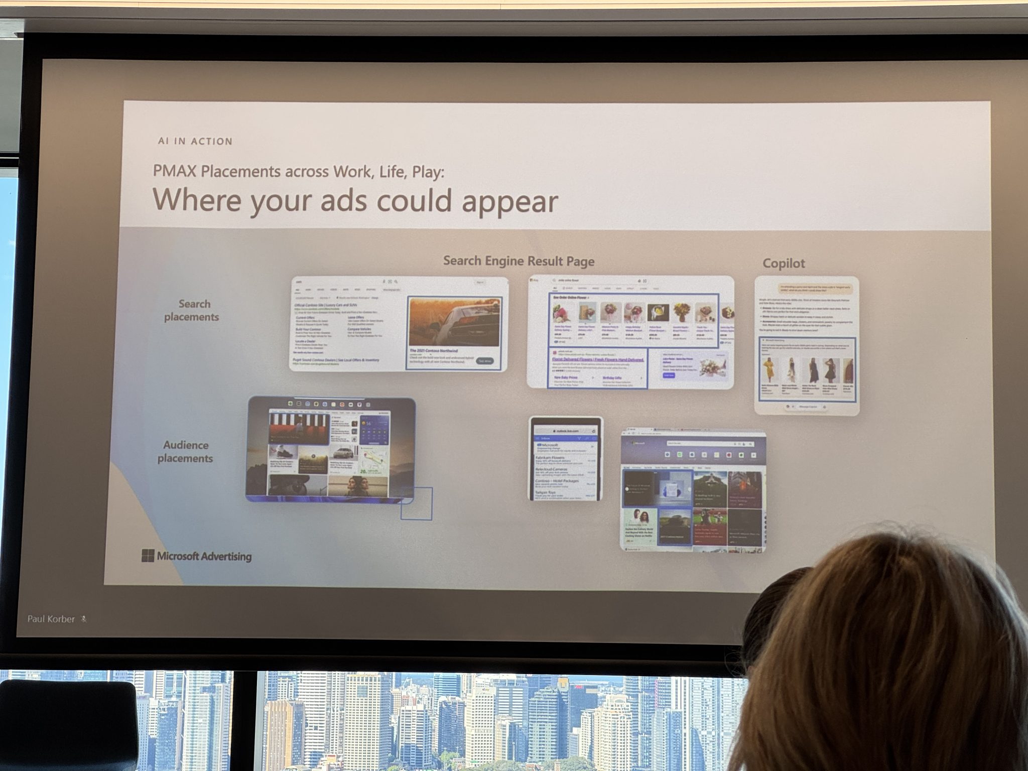 A presentation slide showing examples of ad placements on search and audience platforms at Microsoft's Marketing Event, with a city skyline visible through the window in the background, highlighting how AI can boost ROI.