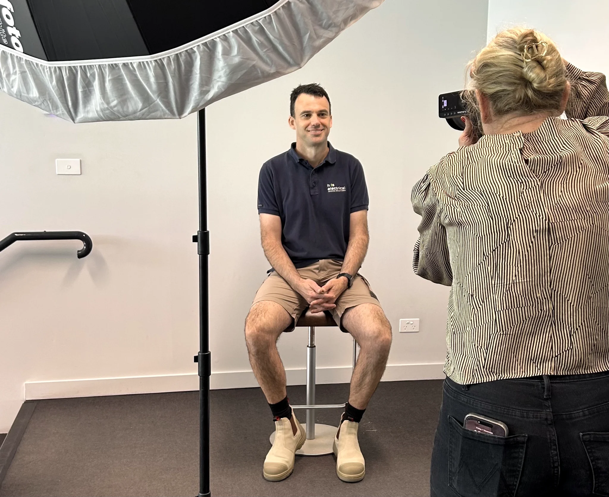 A man sits on a stool in a studio, posing for a Branded Photography session as the photographer captures his image. Studio lighting equipment is visible above.