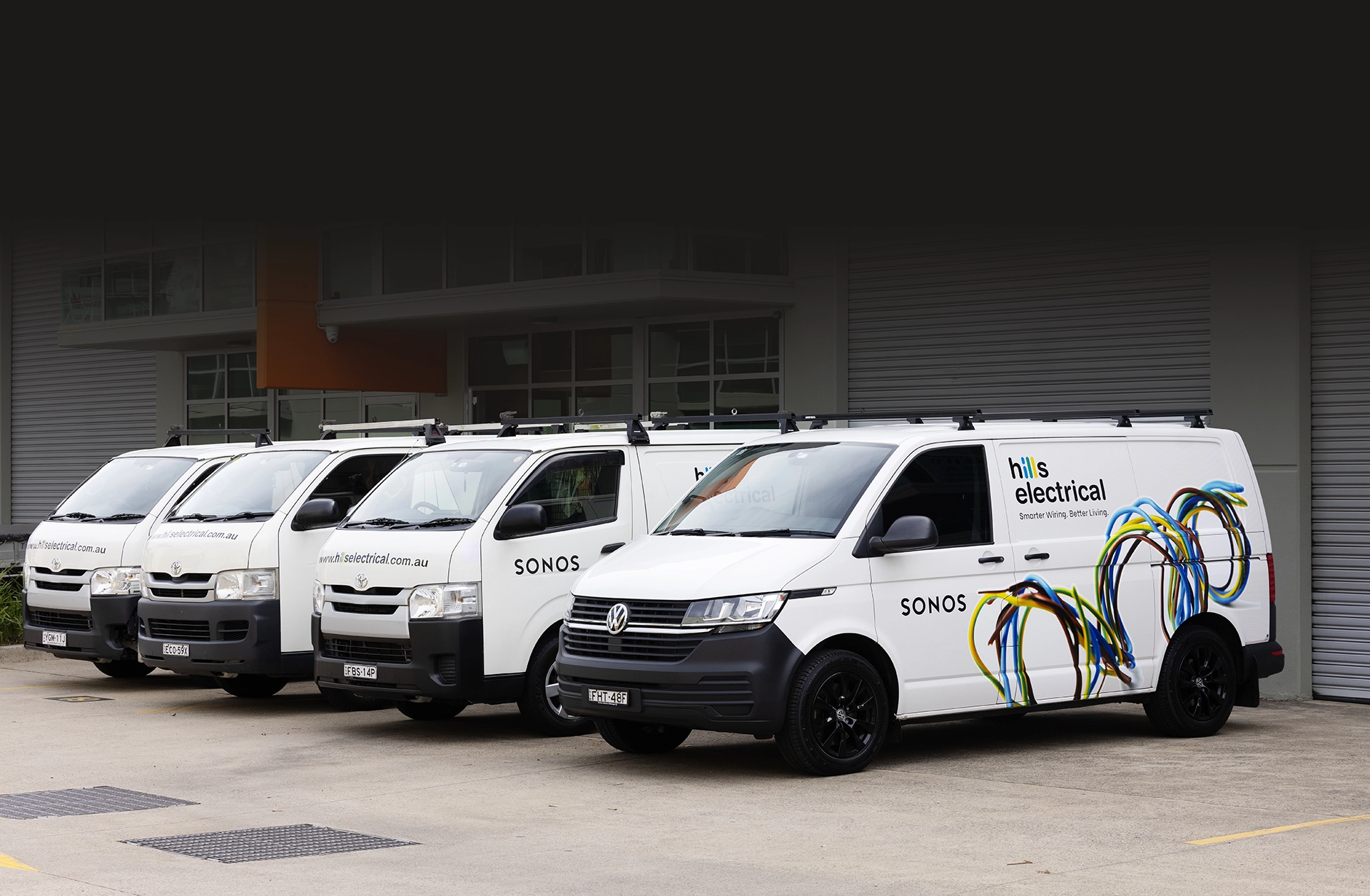 Four white HiLS Electrical service vans with colorful wiring graphics and SONOS branding are parked in a row outside a commercial building with roller doors, showcasing striking branded photography.