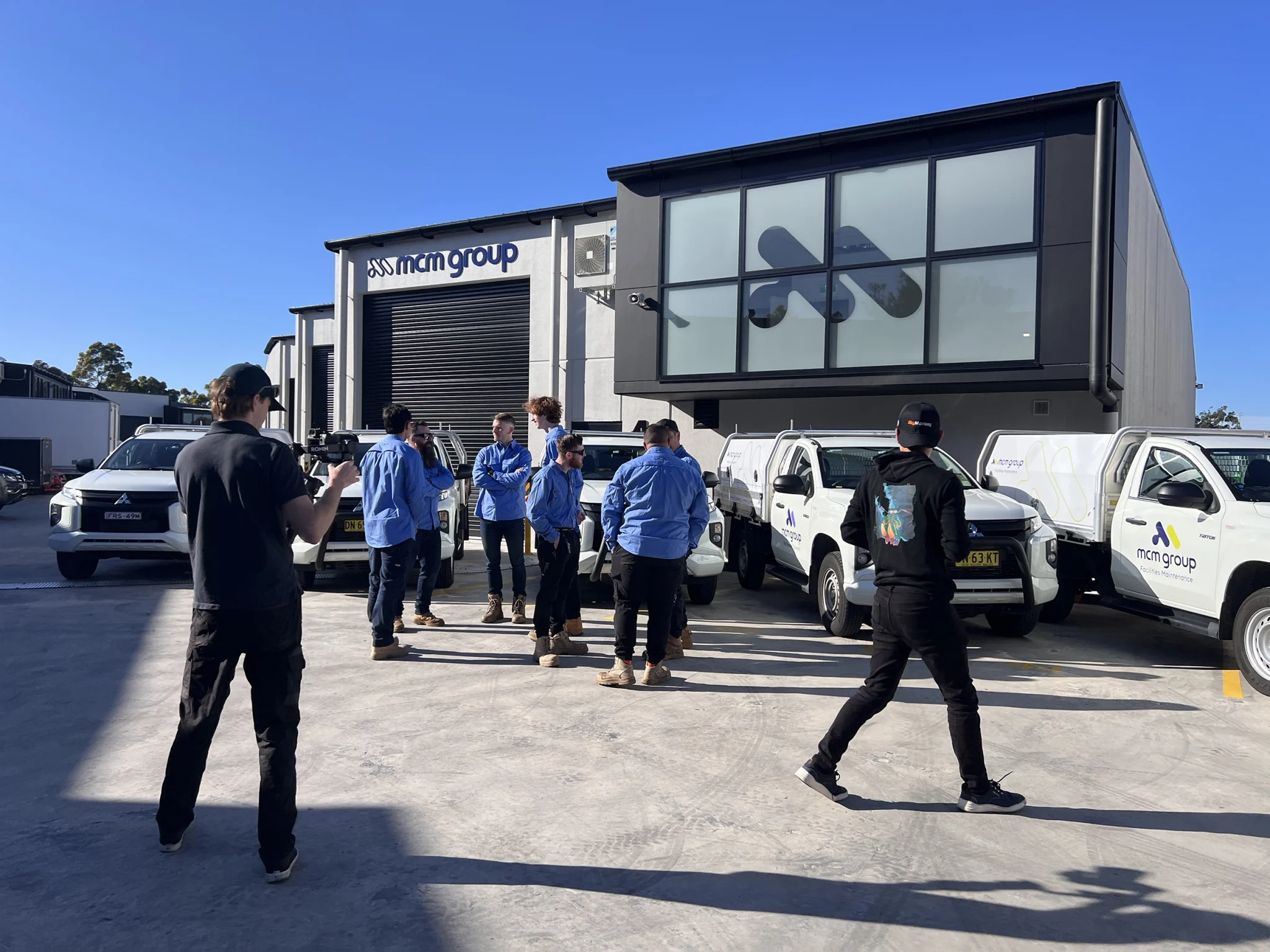 A group of people in work uniforms stand and talk in front of a modern office building, with several company-branded vehicles parked outside—an ideal scene for brand photography.