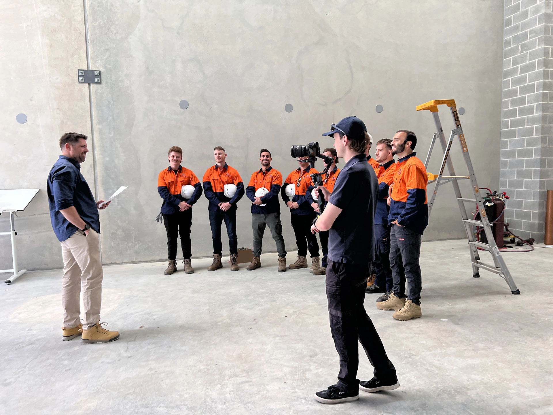 A group of workers in orange shirts stands against a wall holding helmets while a man with papers addresses them and a cameraman films the scene, capturing branded photography in an industrial space.