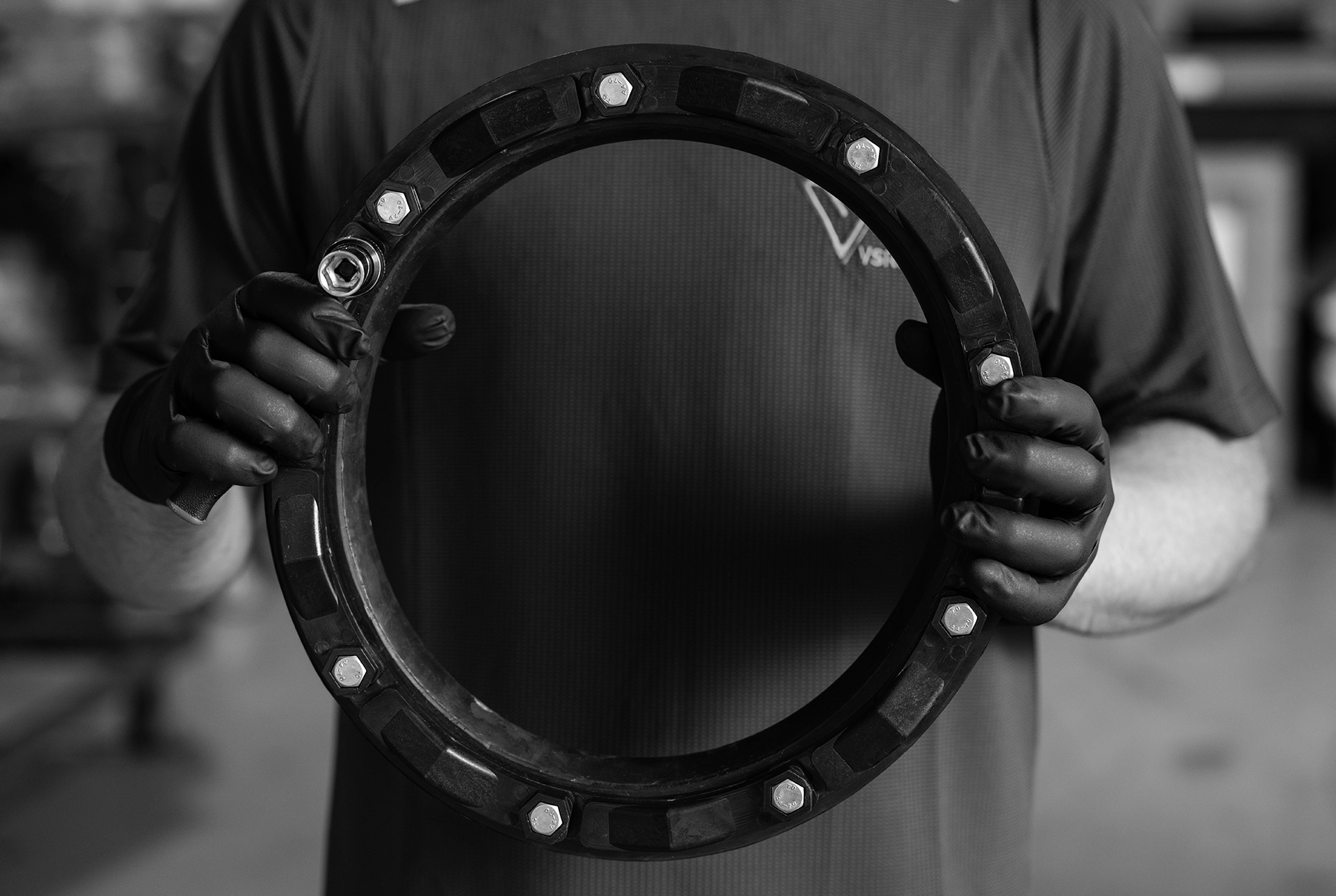 A person wearing gloves holds a large circular metal mechanical part with bolts around its edge, captured in a branded photography style.