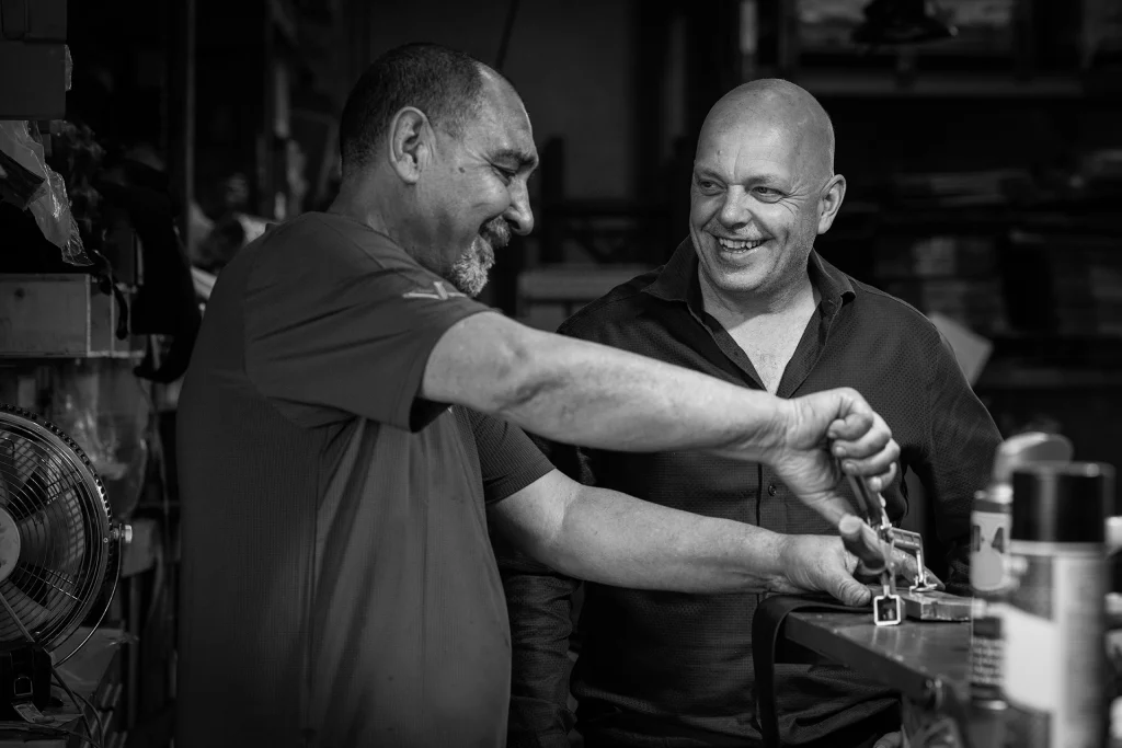 Two men stand at a workbench, one using a wrench on a metal object while the other watches and smiles, captured in a workshop setting emphasizing professional photography.