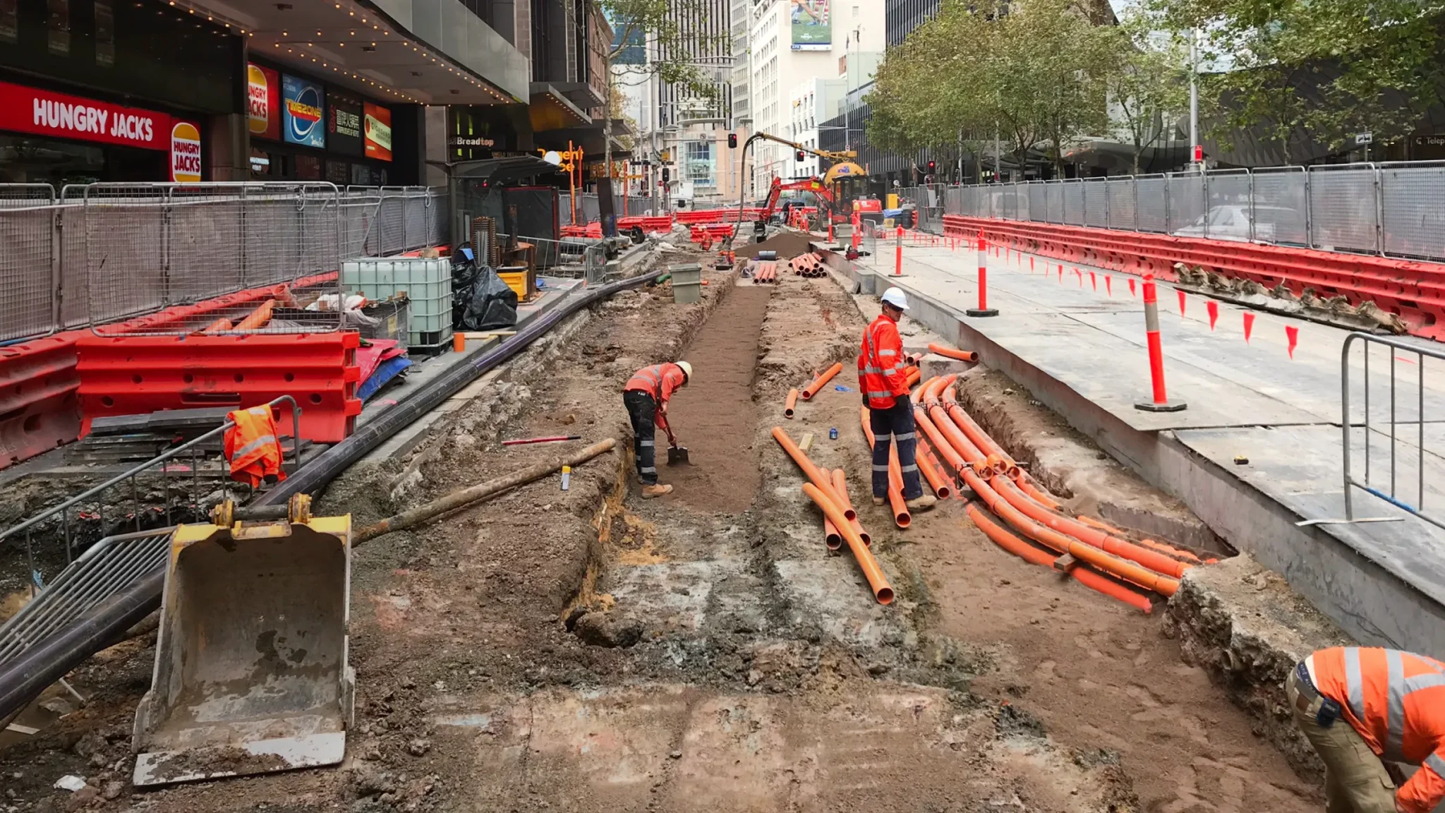 Construction workers in safety gear install orange pipes and work on a partially dug-up city street, surrounded by barriers and equipment—an ideal scene for a construction website highlighting real-world projects of a professional construction company.