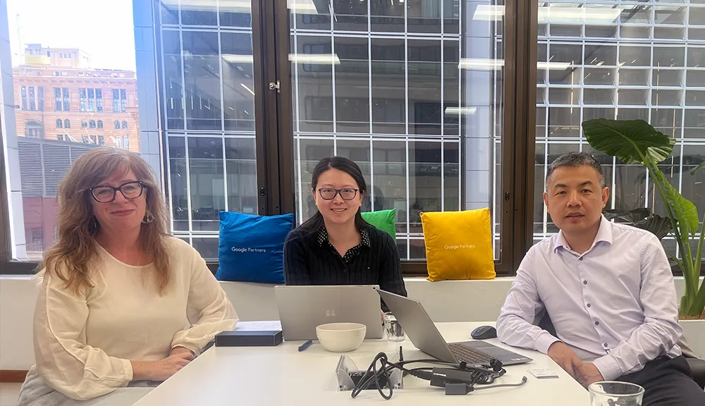 Three people sit at a table in a modern office with large windows, discussing client growth. Two laptops, documents, a bowl, and a power cord are on the table. Colorful pillows decorate the bench behind them.