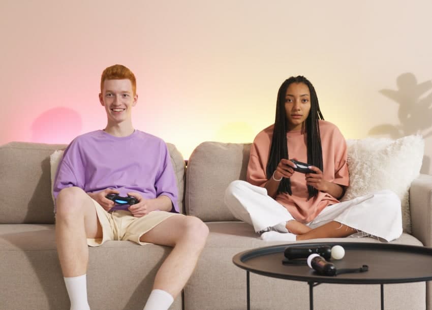Two people sit on a beige couch playing video games. The man on the left is smiling, while the woman on the right looks focused. A round table with objects is in the foreground.